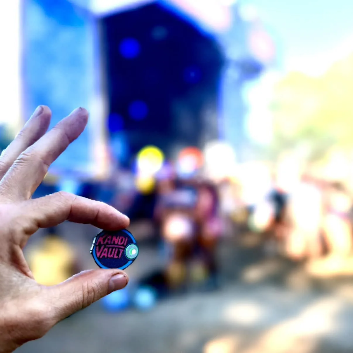 Hand holding a woven KandiVault patch with a blurred festival stage glowing in the background